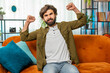 © Andrii Iemelianenko - Happy middle eastern man shouting, celebrating success, winning, goal achievement good news, lottery luck, victory, watching TV championship at modern home apartment. Guy in living room sits on couch