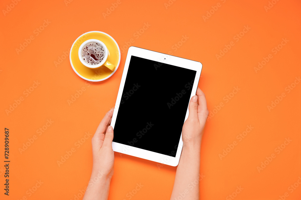 Female hands holding tablet computer with blank screen and cup of coffee on orange background