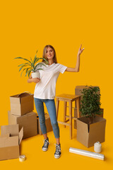 Wall Mural - Young woman with houseplant and cardboard boxes pointing at something on yellow background. Moving house concept