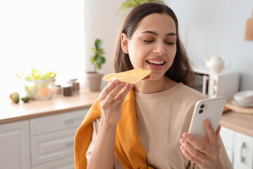 Poster - Young beautiful happy woman with mobile phone eating tasty sandwich in kitchen