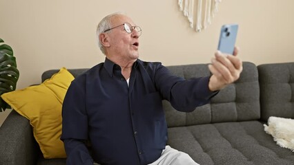 Canvas Print - Joyful senior man sitting on the sofa at home, taking an expressive selfie with a smartphone, smile lighting up the room