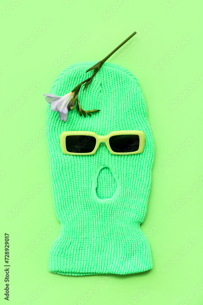 Stylish balaclava with flower and sunglasses on green background