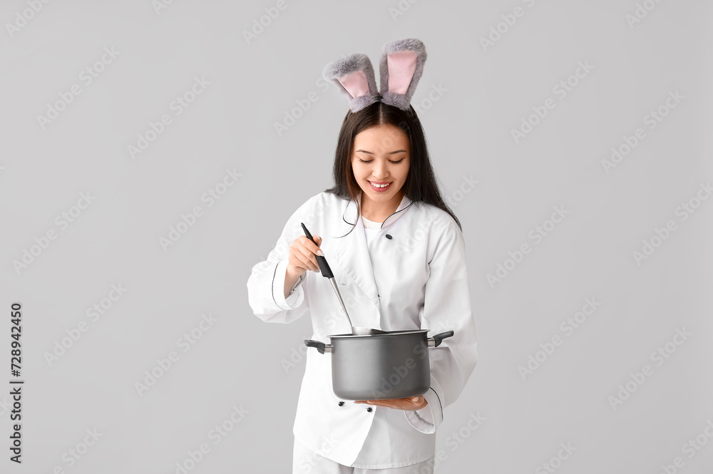 Female Asian chef in bunny ears with cooking pot on light background. Easter celebration