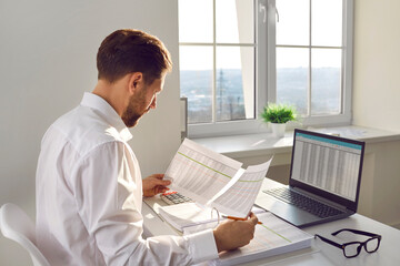 Financial accountant, auditor or business man doing paperwork in the office. Young corporate employee or businessman sitting at desk with laptop computer and looking at data sheets that he is holding