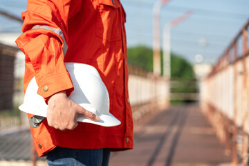 Wall Mural - An engineer or construction worker is holding a white safety helmet, wearing orange coverall, standing on the working platform walkway. Ready to work in the challenge workplace concept scene. 