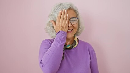 Wall Mural - Confident middle age woman with grey hair, wearing glasses, funny cover of eye with hand, peeking cheerfully over her glasses with a radiant smile. standing against a vibrant isolated pink background.