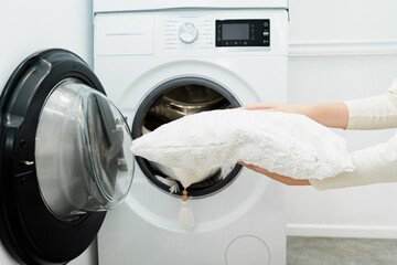 Woman holding clean white pillow in front of the drum of washing machine in laundry room. Washing pillows