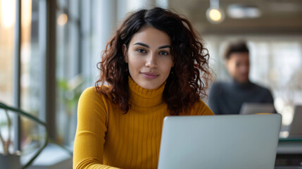 Poster - young woman with red hair, wearing a yellow sweater, is smiling at the camera while working on her laptop at a modern cafe with a blurred figure in the background