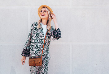 Young beautiful smiling blond hipster woman in trendy summer hippie costume clothes. Carefree female posing in the street at sunny day. Positive model outdoors at sunset. Cheerful and happy in hat