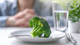Fototapeta Mapy - close-up of a single broccoli floret on a white plate, with a fork beside it, a glass of water in the background, and a person's hands folded in the background
