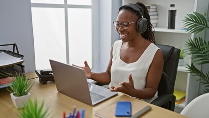 Wall Mural - A cheerful woman with headphones works on a laptop in a modern office, suggesting productivity and technology use.