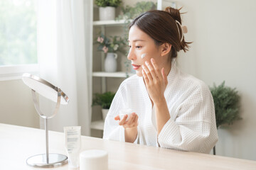 Wall Mural - Facial beauty skin care, smile of pretty asian young woman in bathrobe looking at mirror, hand applying moisturizer lotion on her face, holding jar of skin cream before makeup cosmetic routine at home