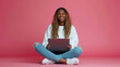 © Studio Nova - cheerful young woman sitting with her legs crossed, holding a laptop on her lap, wearing a white sweatshirt and ripped blue jeans, against a coral pink background