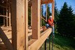 © anatoliy_gleb - Carpenter constructing wooden frame two-story house near the forest. Bearded man holding hammer, dressed in protective helmet and orange safety vest. Concept of ecological modern construction.