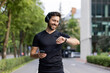 © Tetiana - Smiling young man doing sports and running. Standing outside in the city wearing headphones, holding a mobile phone and looking at a smart watch
