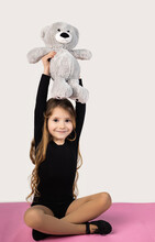 Vertical Shot Of A Little Gymnast Girl On A Yoga Mat Wearing A Black Suit, Holding Her Hands Up With A Teddy Bear.