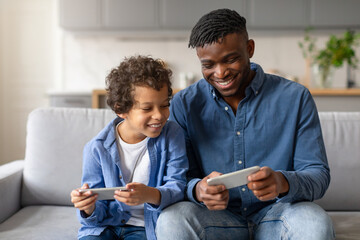 Wall Mural - Joyful black father and son playing video games on cellphones together