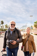 Wall Mural - Vertical smiling Caucasian mature tourist couple standing posing looking at camera in street with their luggage. Husband and wife enjoying their retirees holidays wearing winter clothes on sunny day