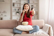 © Prostock-studio - Cheerful young asian woman eating popcorn and holding remote control
