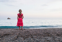 Young Girl Standing On The Beach Looking Out Onto The Ocean Wearing Red Dress. Elba Island, Italy