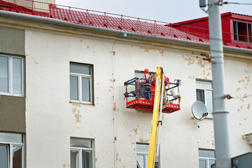 Wall Mural - Workers repairing wall facade, painting and plastering old building. Construction workers working in lifting bucket. Plaster work, men repair cracks on facade. Men repair window jamb, external work.