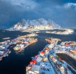 Wall Mural - Aerial view of snowy Henningsvaer fishing village, Lofoten islands, Norway in winter at sunset. Top drone view of mountains in snow, sea, boats, bridge, road, cloudy sky, town, rorbuer and houses