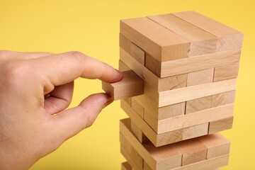 Sticker - Playing Jenga. Man removing wooden block from tower on yellow background, closeup