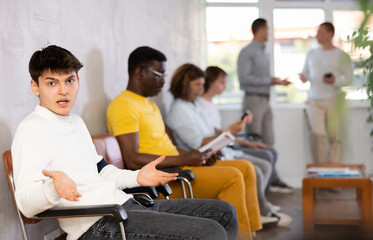 Wall Mural - Disgruntled young guy sitting on the bench of visitors in hospital waiting for his turn for consultation