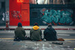 Three homeless individuals seated on a sidewalk with their backs to the camera, facing a colorful graffiti wall