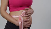 Close-up. A Pregnant Woman With A Pink Ribbon On Her Belly Stands On A White Background. A Woman Gently Hugs Her Belly With Both Hands