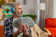 © Andrii Iemelianenko - Happy excited rich joyful senior man in wireless headphones showing wasting money cash around, more tips earnings, big profit, win lottery jackpot share celebrating success on sofa at home. Copy-space