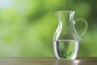 Glass jug with clear water on wooden table against blurred green background, closeup. Space for text