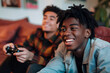 black brown young men playing video game smiling joystick in hands teenagers friends friendship happy complicity sofa in a living room having fun wearing casual shirts denim dreadlocks cheerful upbeat