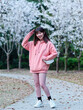 Beautiful young Chinese woman smiling under blooming cherry tree, wearing pink loose oversized top with small white shoulder bag. Candid moment. Emotions, people, beauty, youth and lifestyle portrait.