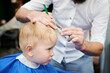 Man barber cutting little boy's hair using comb and scissors. Child getting haircut from adult male, likely barber. Professional hairdresser and cute client at modern barbershop.
