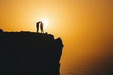 Silhouettes Of Pair Standing On Cliff, Sunrise In Background