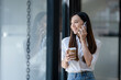 © Wasana - A cheerful young businesswoman engaging in a phone conversation while holding a coffee cup, inside a bright modern office space..
