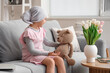 Little girl after chemotherapy with toy bear sitting on sofa at home. International Childhood Cancer Day