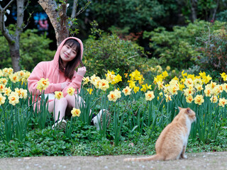 Beautiful young Chinese woman sitting in yellow daffodil flower field smiling with homeless ginger tabby cat. Candid happy moment. Emotions, people, beauty, youth and lifestyle portrait.