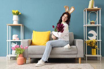 Sticker - Young woman in bunny ears with tulips sitting on sofa at home. Easter celebration