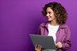 A woman stands in front of a vibrant purple wall while holding a laptop computer.
