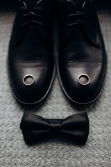 groom's men's accessories black leather shoes, wedding rings and a bow tie on a black table