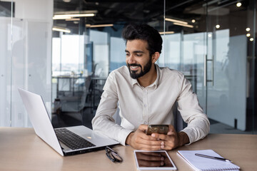 Smiling young Indian male businessman working in the office, sitting at a desk, holding a mobile phone and talking on a video call through a laptop
