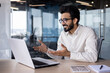 © Tetiana - Online meeting, conference. Indian young man, office worker and businessman prays on laptop for video call, sitting in office at desk, gesturing with hands and smiling