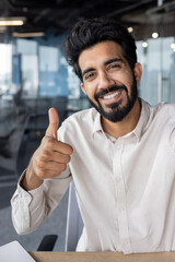 Wall Mural - Close-up portrait of a young Indian man talking on the phone through a video call. He smiles and shows the super sign with his finger on the camera. Vertical photo