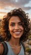© Tom - Portrait of a young woman with curly brunette hair, smiling in a natural outdoor setting during sunset, wearing a casual tank top and backpack.