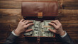 Hands of a person in a business suit are counting US dollar bills from a leather briefcase on a wooden table.