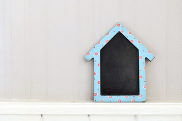 A blank chalkboard in the shape of a house, set on a pale grey wallpaper background. The board is rustic and slightly worn adding to its charm.