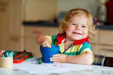 Wall Mural - Cute adorable baby girl learning painting with water colors. Little toddler child drawing at home, using colorful brushes. Healthy happy daughter experimenting with colors, water at home or nursery.