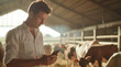 A focused farmer in a white shirt uses a smartphone with a herd of cows in the background inside a barn